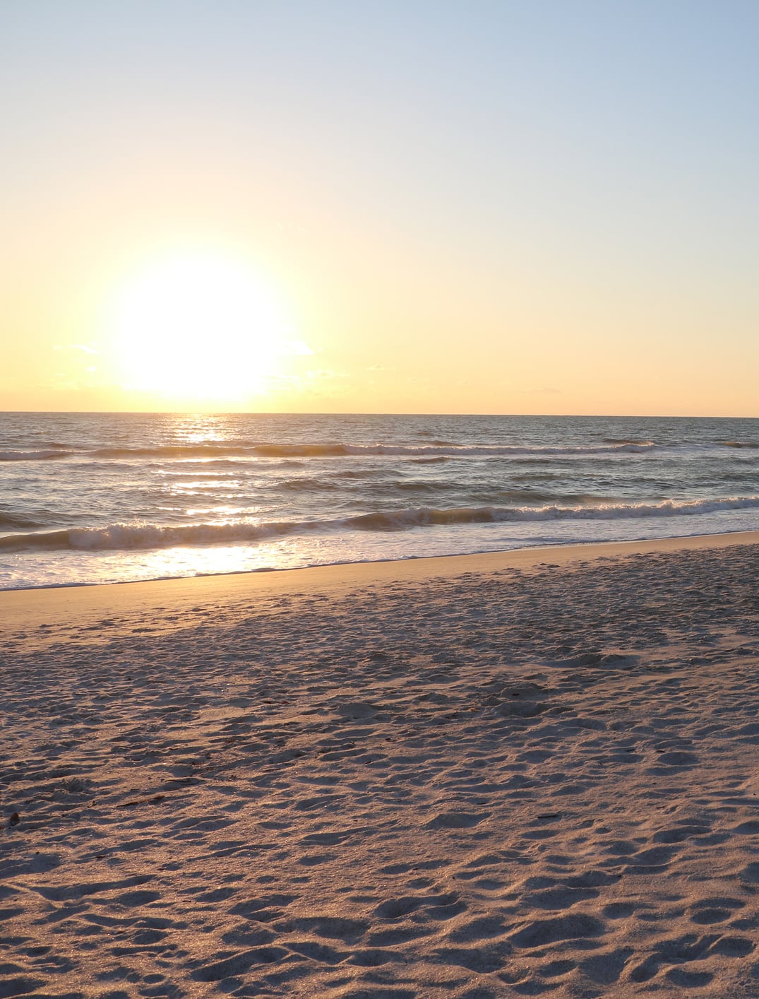 A bright sun sets over the ocean, casting a golden glow on the breaking waves and stretching across the wide, textured sandy beach.