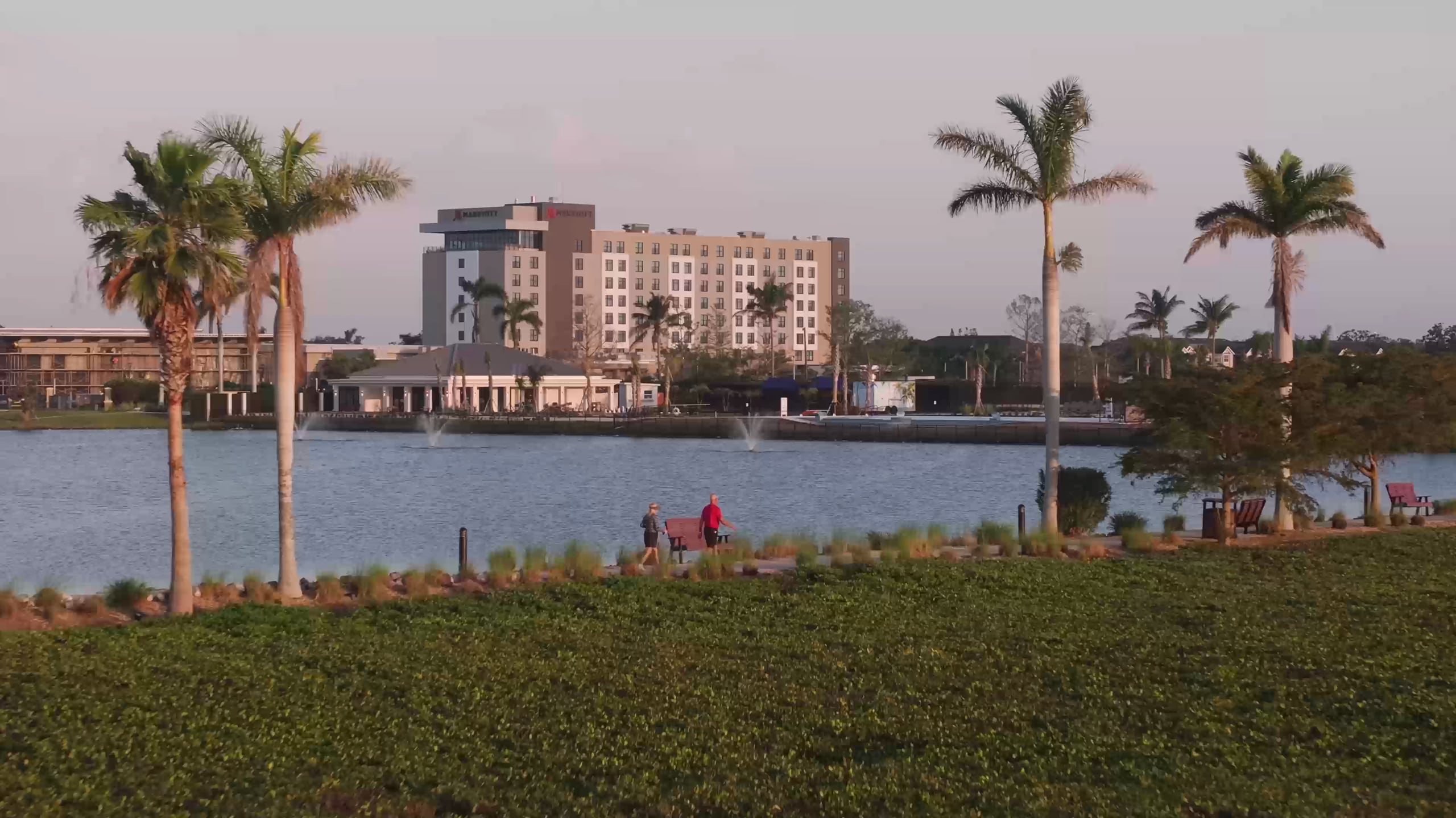 Two people walk along a path beside a lake with fountains and palm trees. A distant "MARRIOTT" hotel overlooks the water.