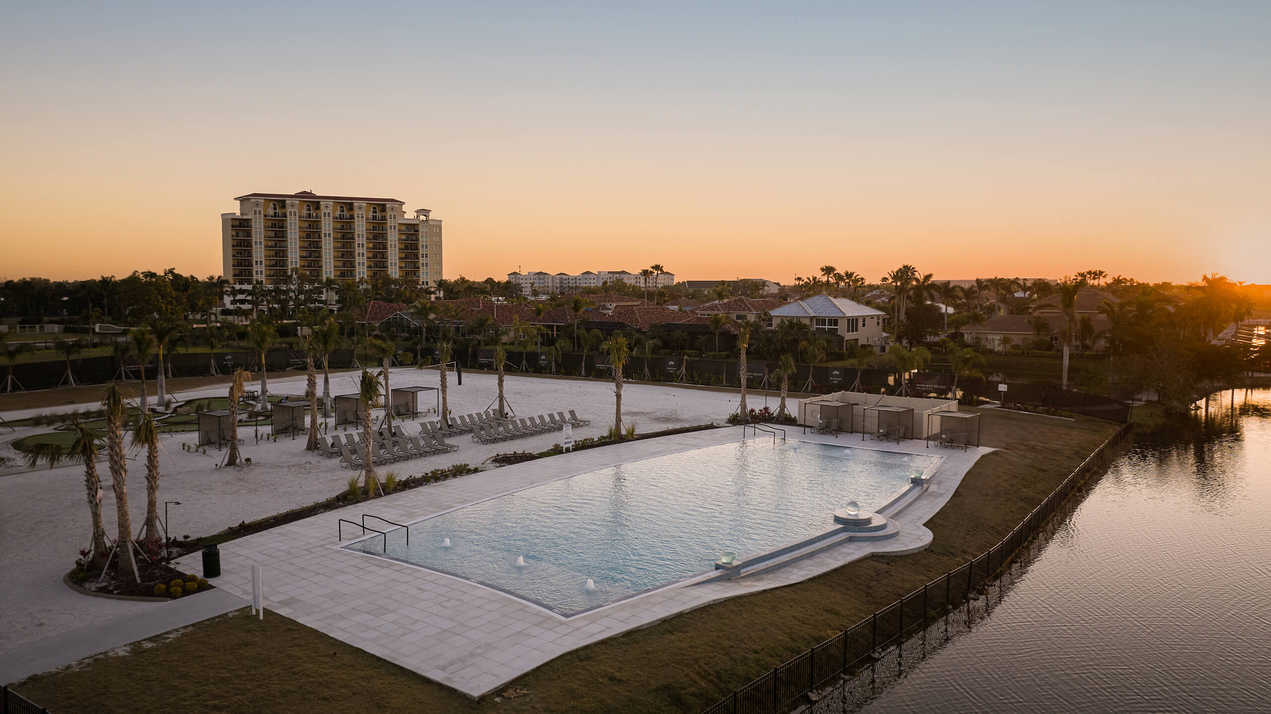 A large resort-style swimming pool reflects the orange sunset, flanked by lounge chairs and palm trees. A multi-story building and a lake are visible in the background.
