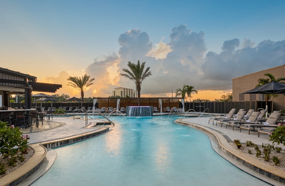Resort pool with waterfall and fountains surrounded by lounge chairs, a bar, and palm trees at sunset. Depths: "1 FT 6 IN", "0.5 M", "3 FT 3 IN", "1 M".