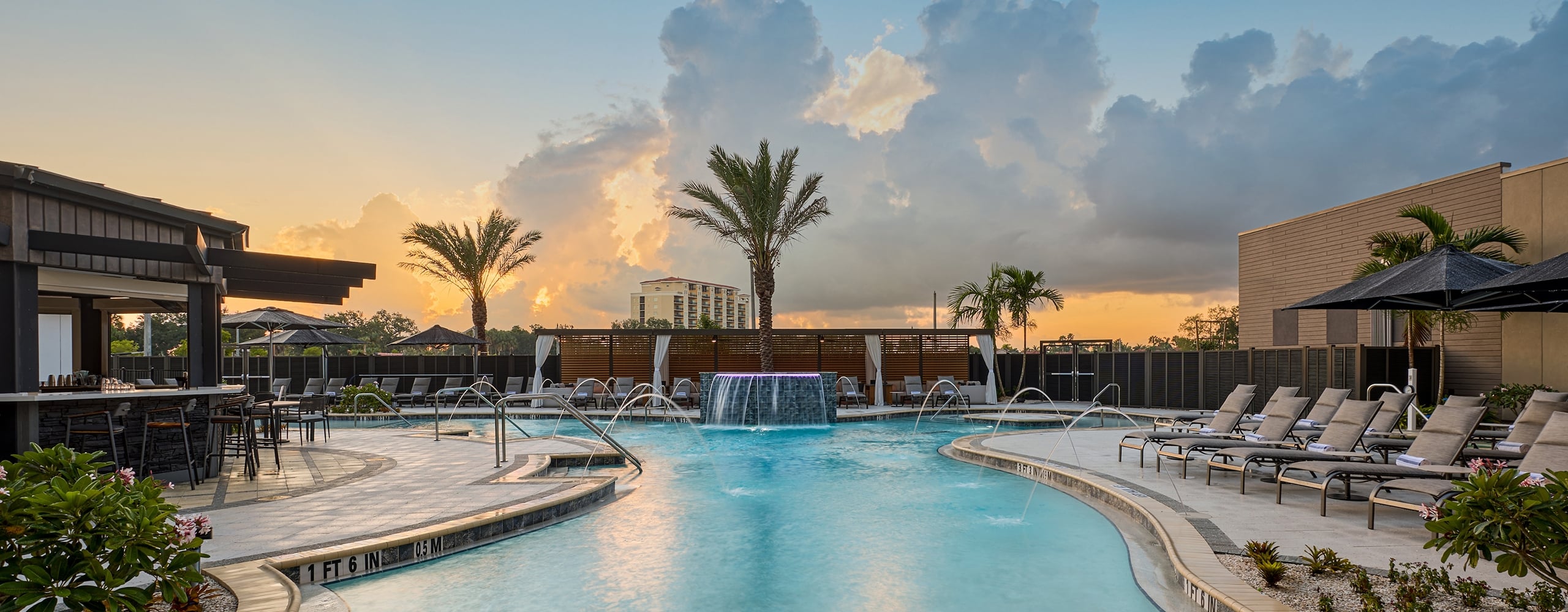 Resort pool with waterfall and fountains surrounded by lounge chairs, a bar, and palm trees at sunset. Depths: "1 FT 6 IN", "0.5 M", "3 FT 3 IN", "1 M".