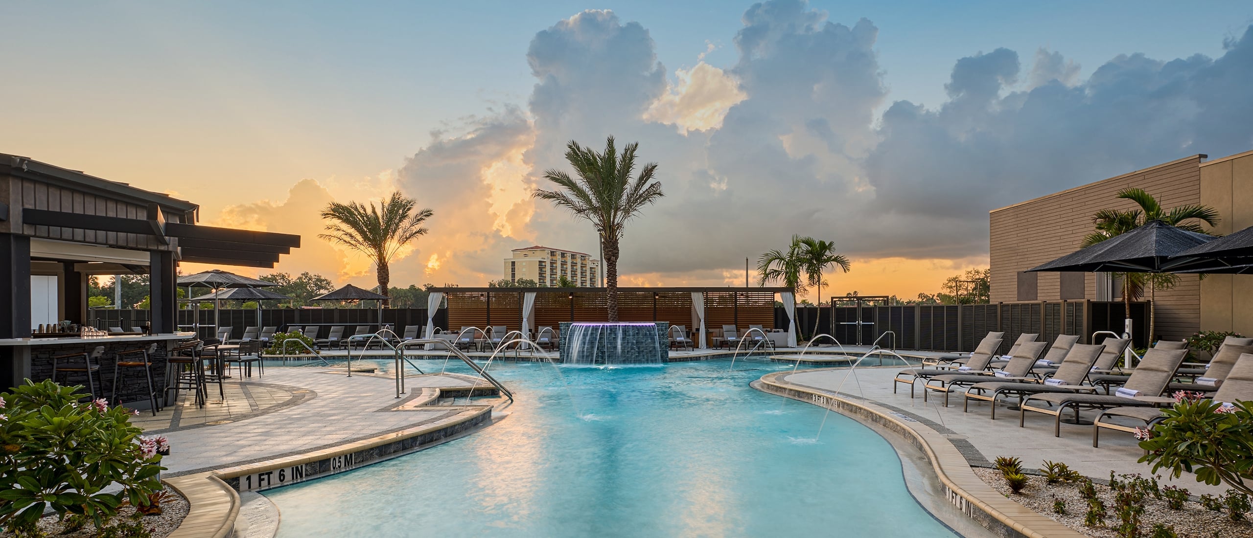 Resort pool with waterfall and fountains surrounded by lounge chairs, a bar, and palm trees at sunset. Depths: "1 FT 6 IN", "0.5 M", "3 FT 3 IN", "1 M".