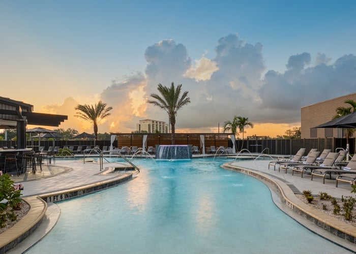 Resort pool with waterfall and fountains surrounded by lounge chairs, a bar, and palm trees at sunset. Depths: "1 FT 6 IN", "0.5 M", "3 FT 3 IN", "1 M".