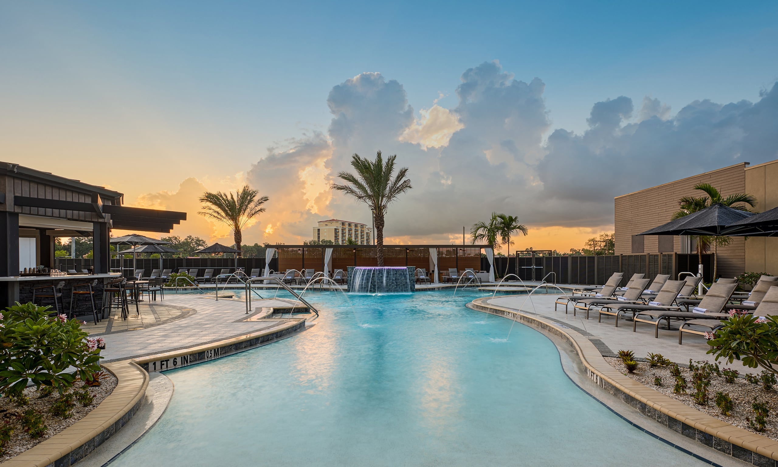 Resort pool with waterfall and fountains surrounded by lounge chairs, a bar, and palm trees at sunset. Depths: "1 FT 6 IN", "0.5 M", "3 FT 3 IN", "1 M".