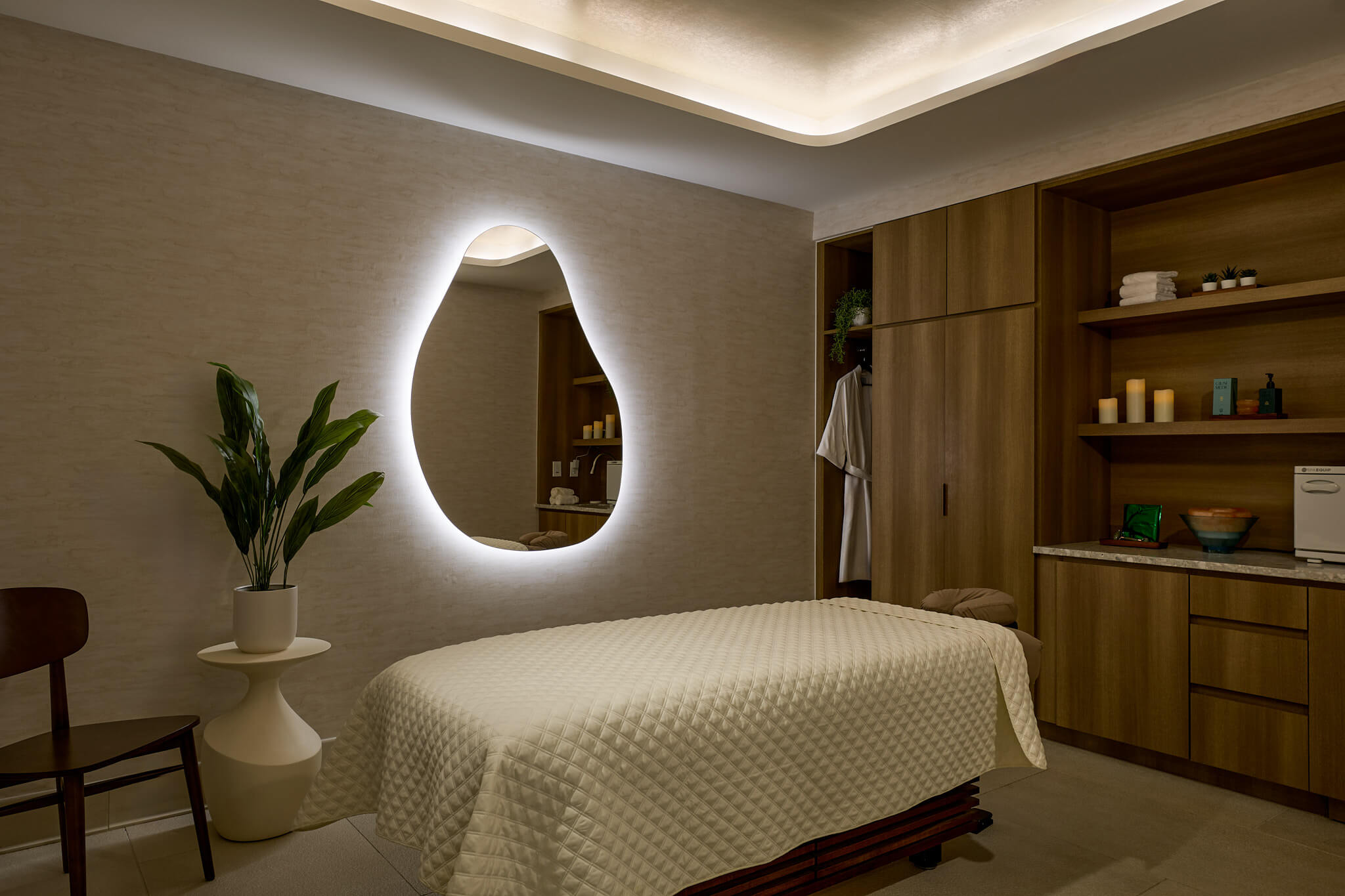 A massage table covered in white is centrally positioned in a softly lit spa room with a glowing organic-shaped mirror, a potted plant, and wood cabinets with shelves.