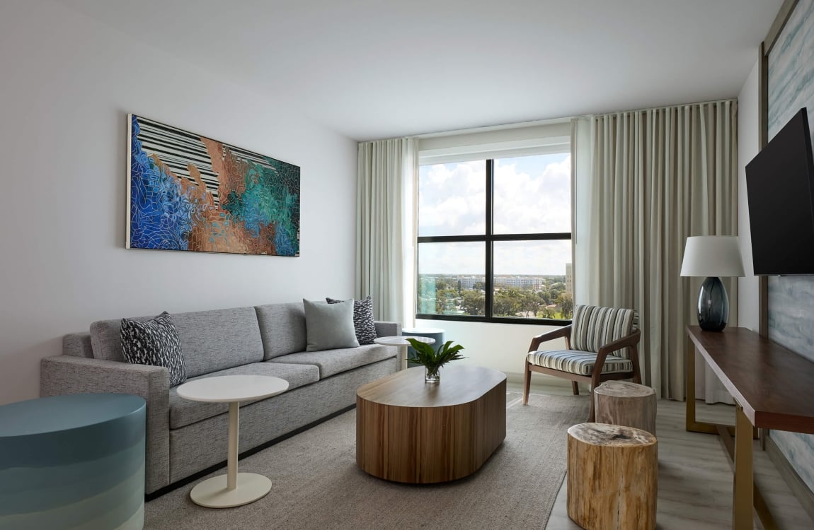 A modern living room shows a gray sofa centered with an oval coffee table. A large window offers city views, while abstract art hangs above the sofa and a striped armchair sits nearby.