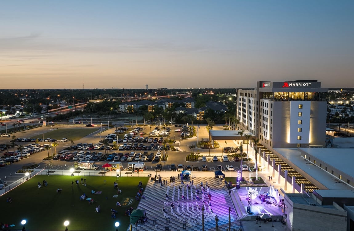 An illuminated Marriott hotel overlooks an active outdoor evening event with a stage and people on a lawn, amidst a busy cityscape at dusk.