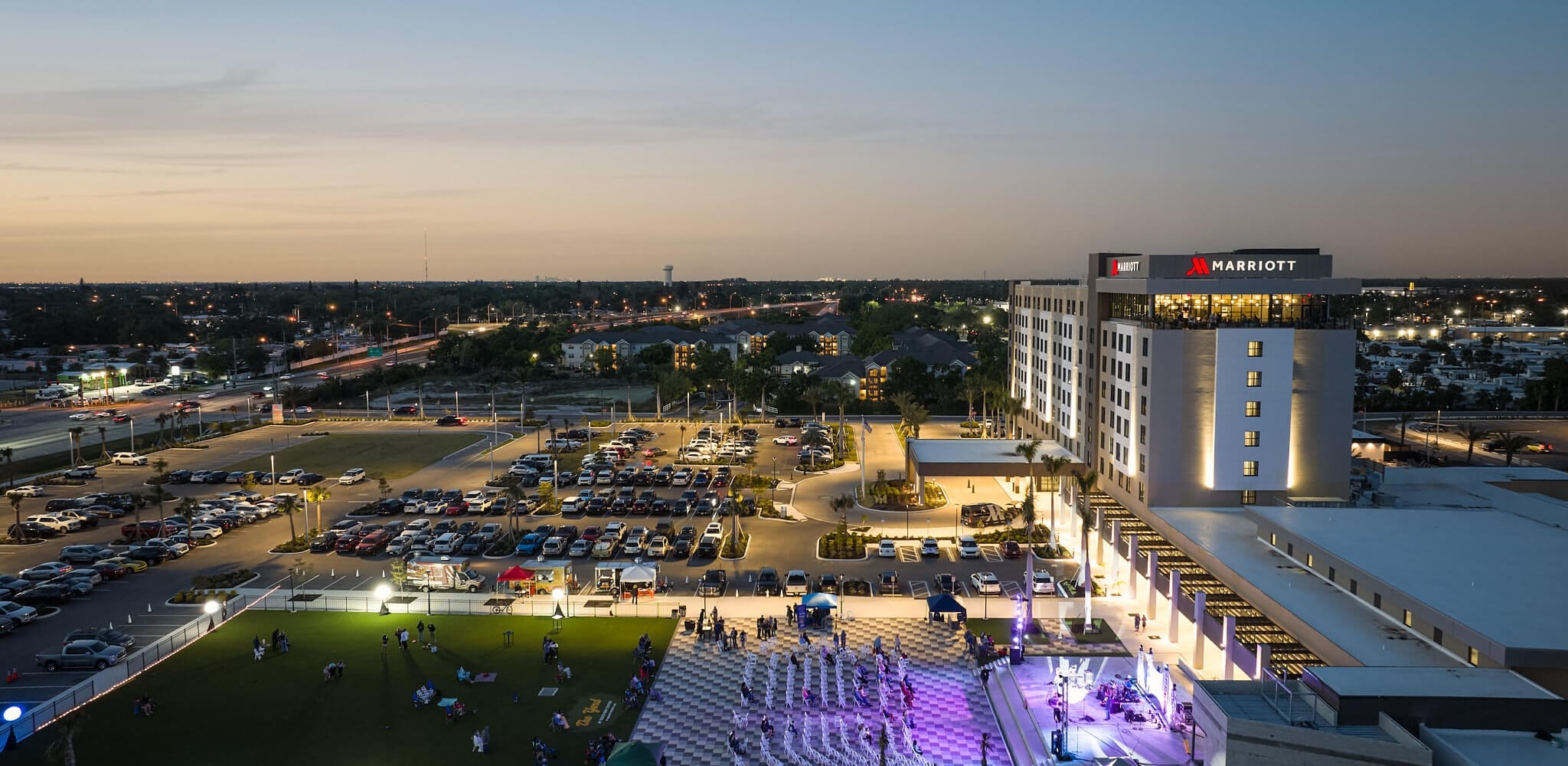 An illuminated Marriott hotel overlooks an active outdoor evening event with a stage and people on a lawn, amidst a busy cityscape at dusk.