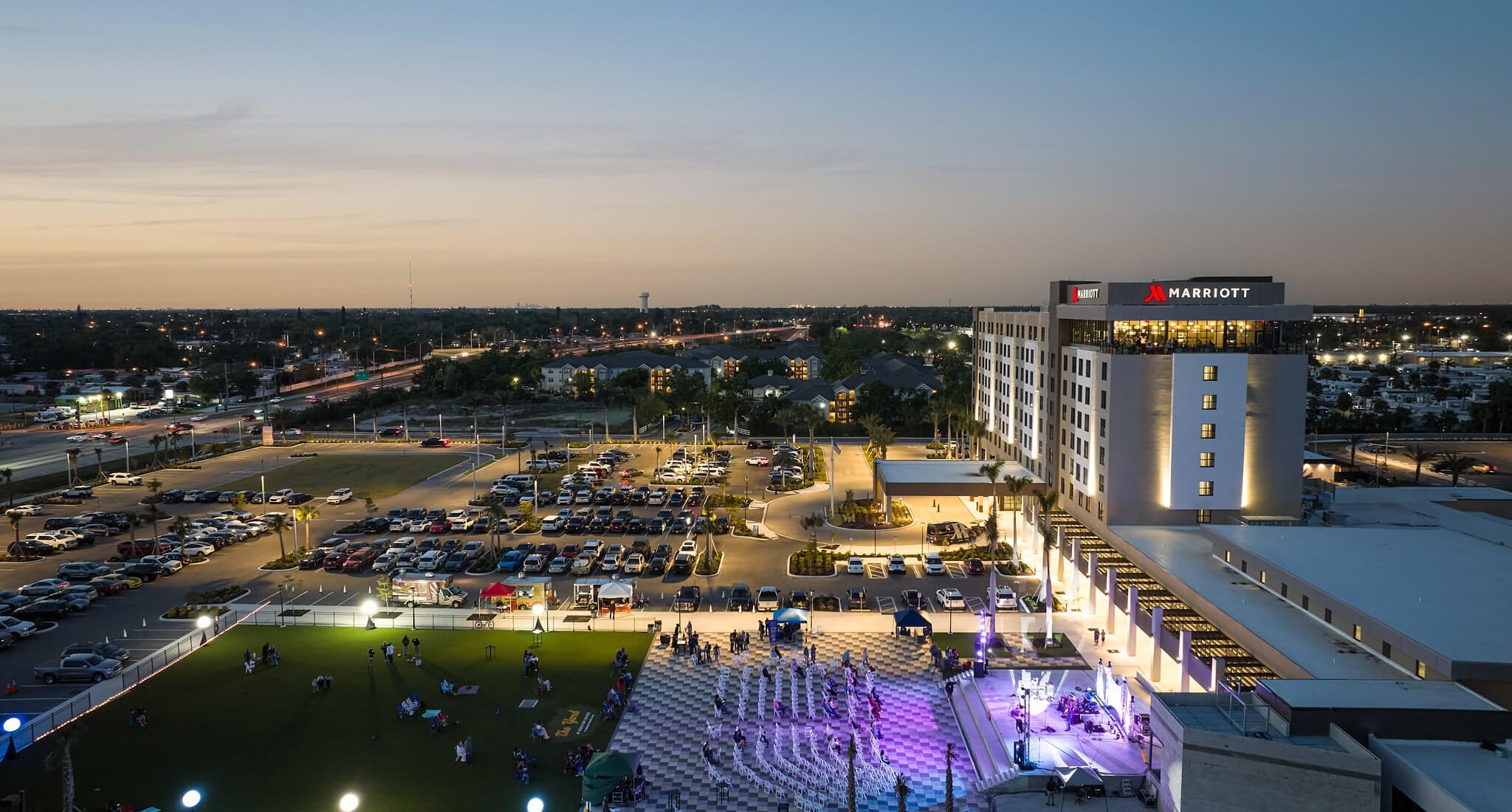 An illuminated Marriott hotel overlooks an active outdoor evening event with a stage and people on a lawn, amidst a busy cityscape at dusk.