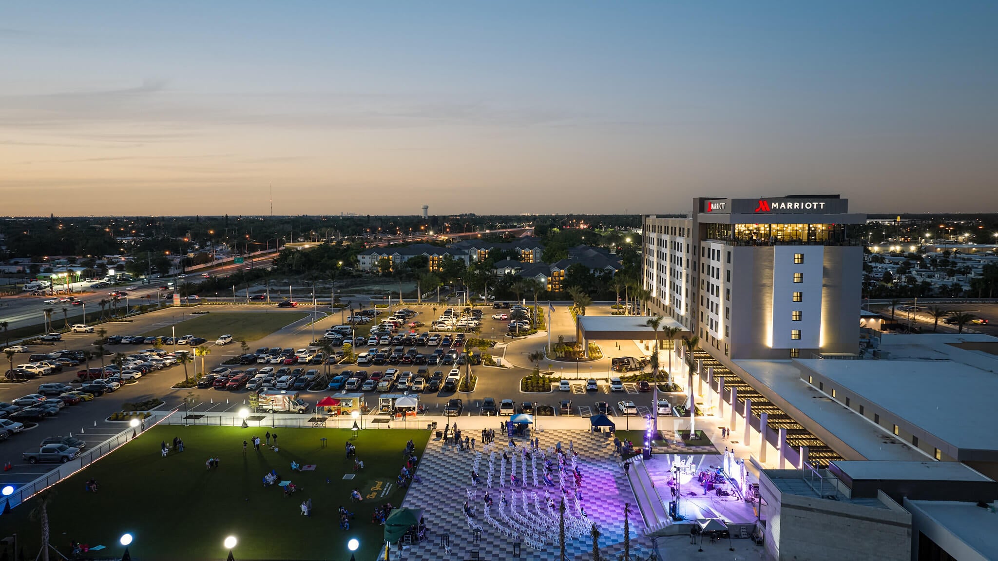 An illuminated Marriott hotel overlooks an active outdoor evening event with a stage and people on a lawn, amidst a busy cityscape at dusk.