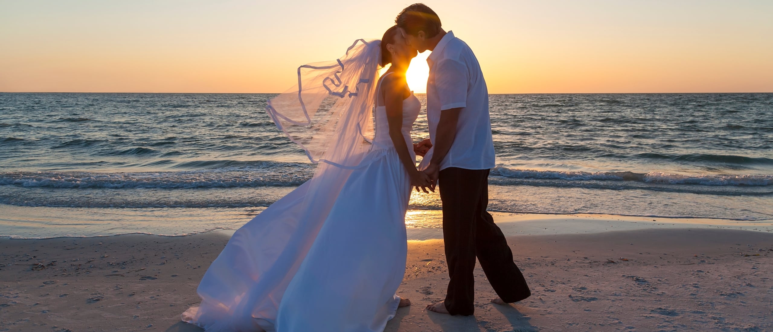 Bride and groom holding hands and kissing on a beach at sunset with the ocean waves in the background.