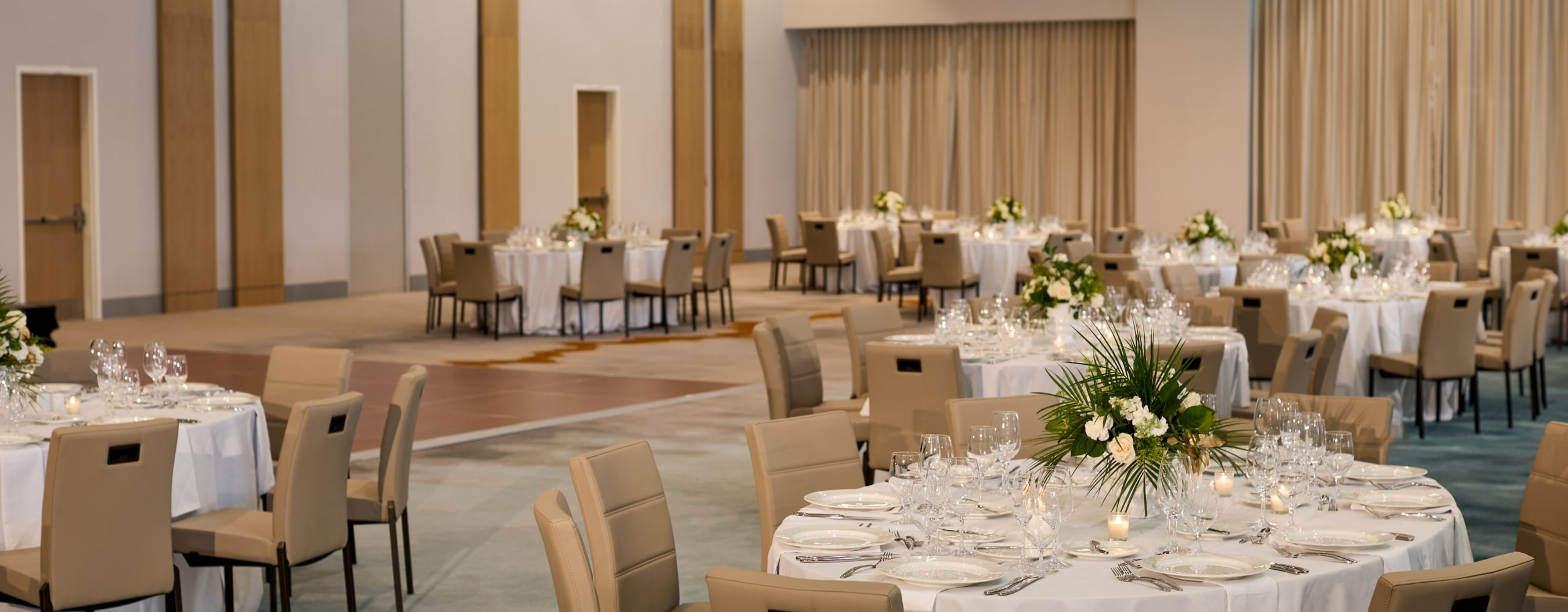 A large, elegantly set ballroom features multiple round tables draped in white linen with floral centerpieces and surrounded by tan upholstered chairs.