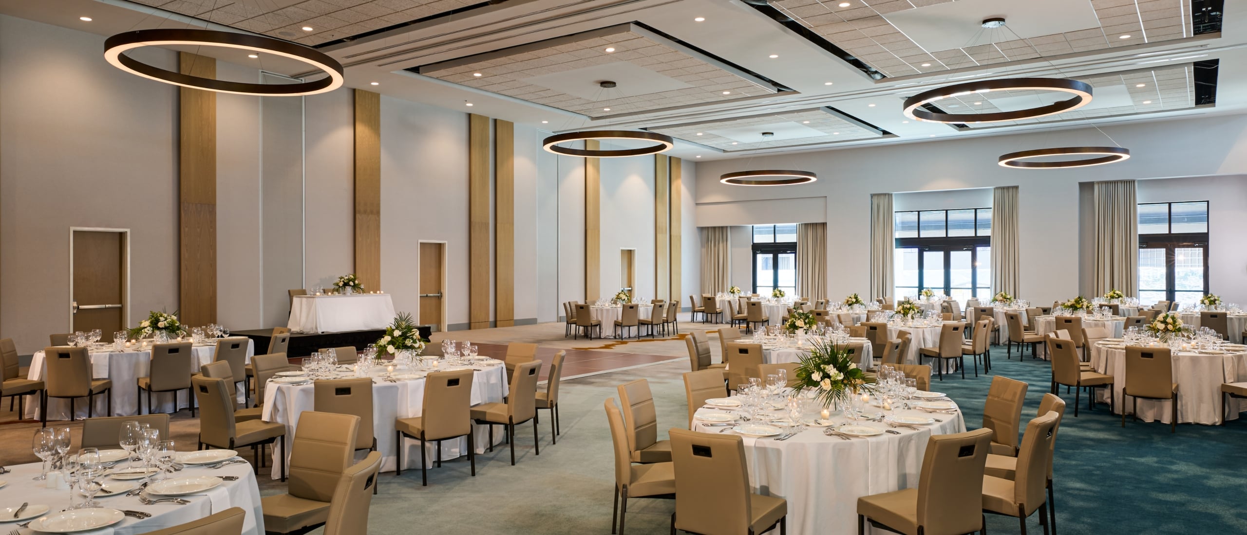 Spacious, modern ballroom set for a banquet with numerous round tables draped in white linen, tan chairs, and illuminated by large circular ceiling light fixtures.