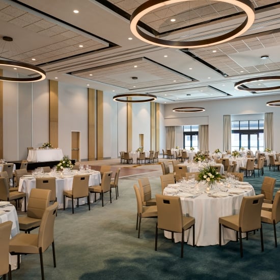 Spacious modern ballroom set for a banquet with numerous round tables, white linens, tan chairs, and distinctive circular overhead light fixtures.