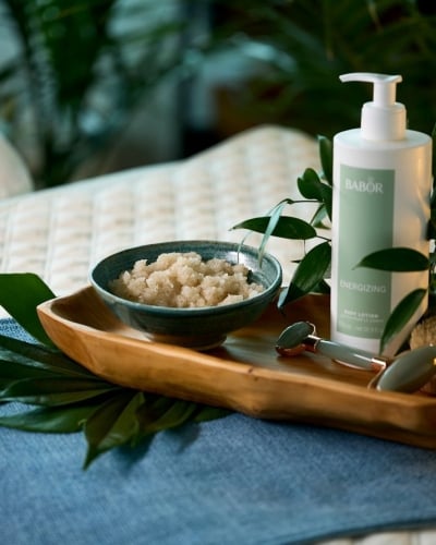A wooden tray holds a BABOR body lotion, a bowl of scrub, a jade roller, and a body brush, set among green foliage on a blue runner over a quilted surface.