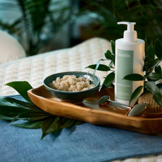 A wooden tray holds a BABOR body lotion, a bowl of scrub, a jade roller, and a body brush, set among green foliage on a blue runner over a quilted surface.