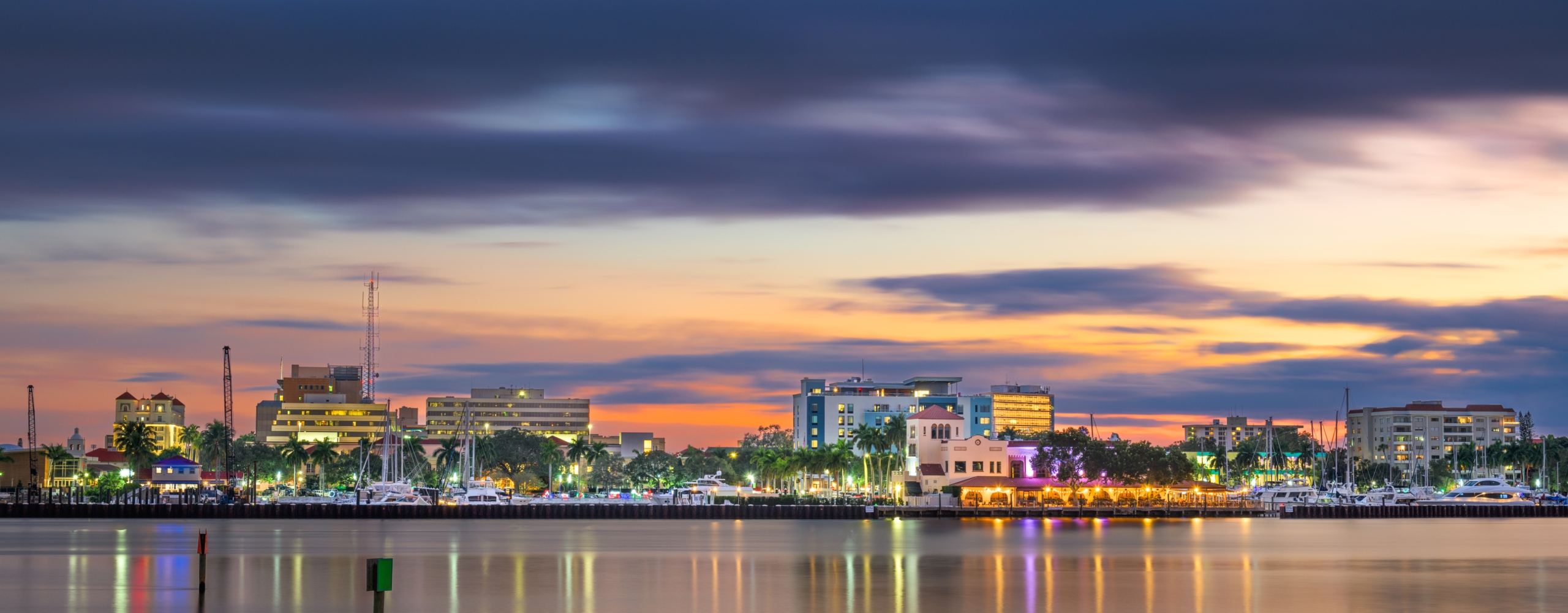 City skyline with illuminated buildings reflecting on calm water under a dramatic twilight sky.