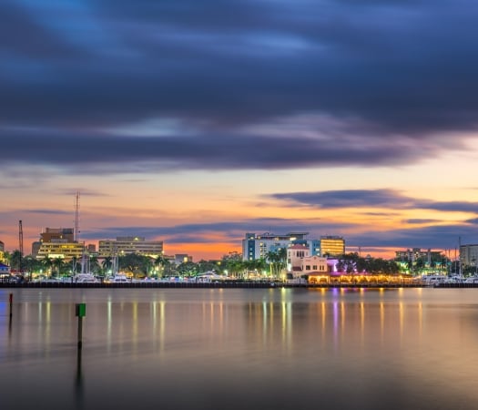 City skyline with illuminated buildings reflecting on calm water under a dramatic twilight sky.