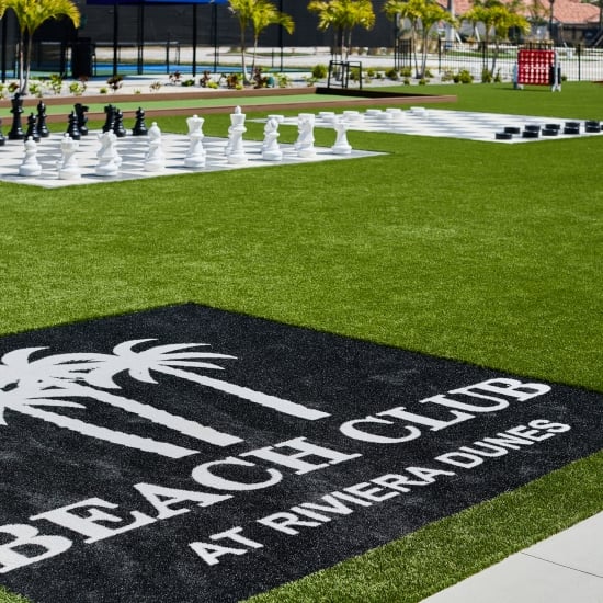 Outdoor recreation area featuring a large chess set, checkerboard, and a black mat reading "Beach Club at Riviera Dunes" on bright green artificial turf.