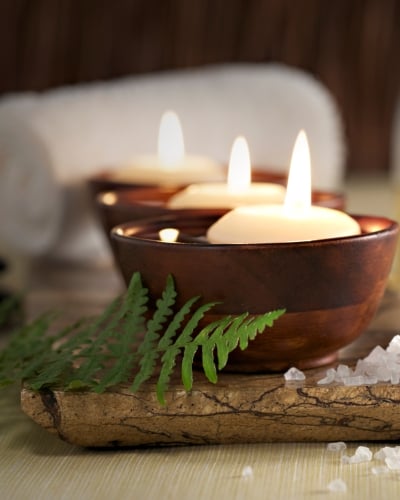 Floating candles in dark wooden bowls are arranged on a surface with stacked smooth black stones, a green fern frond, coarse white salt crystals, and a bottle of oil in the background.