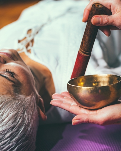 Close-up overhead view of a woman receiving sound healing using a small brass singing bowl held in another person's hand near her head.
