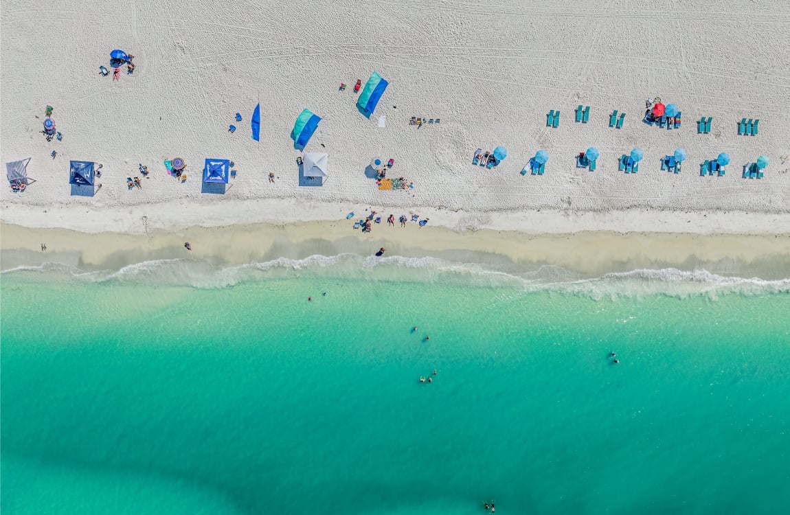 Aerial view of bright white sand beach meeting brilliant turquoise ocean water with scattered beach umbrellas and sunbathers.