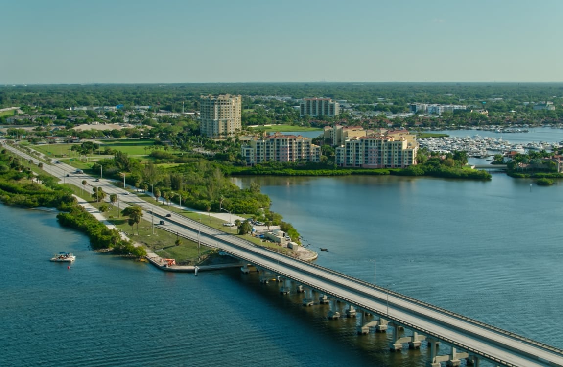 Aerial view of a causeway bridge crossing blue water, connecting to a lush, green shoreline featuring a highway, waterfront condominiums, and a marina full of boats.