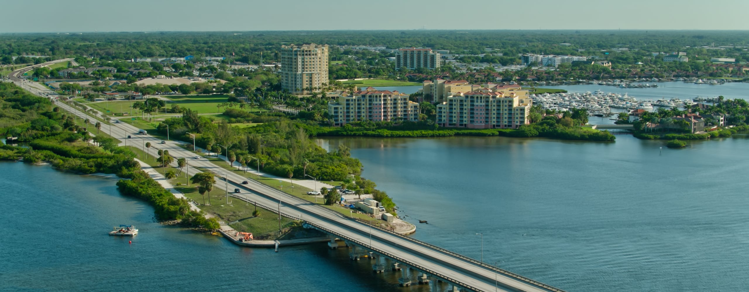 Aerial view of a causeway bridge crossing blue water, connecting to a lush, green shoreline featuring a highway, waterfront condominiums, and a marina full of boats.