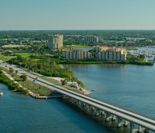 Aerial view of a causeway bridge crossing blue water, connecting to a lush, green shoreline featuring a highway, waterfront condominiums, and a marina full of boats.