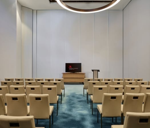 Rows of tan conference chairs face a large screen displaying the Marriott Resort Palmetto logo in an empty meeting room with light walls and a blue patterned carpet.