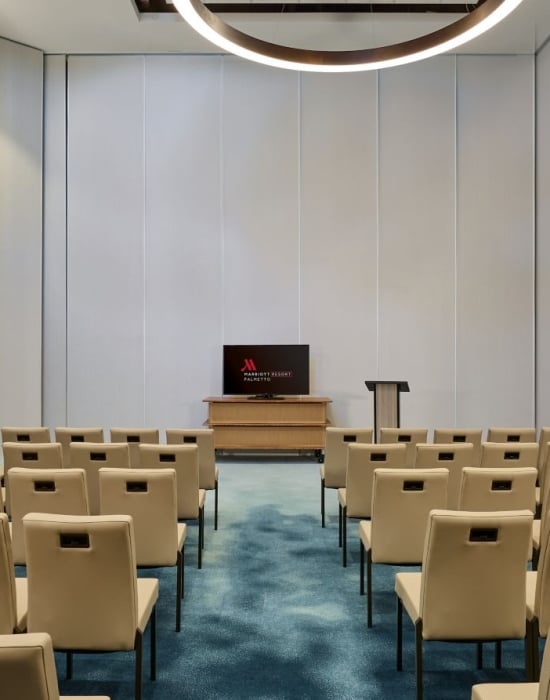 Rows of tan conference chairs face a large screen displaying the Marriott Resort Palmetto logo in an empty meeting room with light walls and a blue patterned carpet.