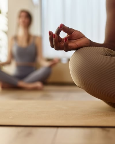 Close-up of a person in yoga attire meditating in the lotus position with another person blurred in the background.