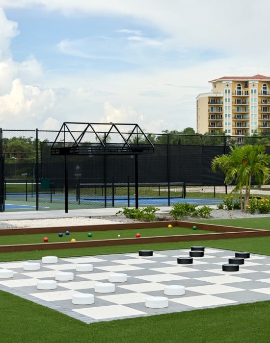 Oversized outdoor checkers board on green turf in front of a fenced tennis court.