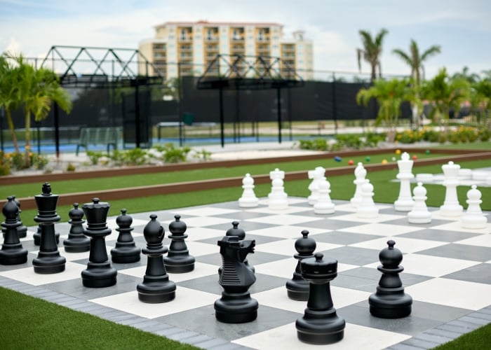Oversized black and white outdoor chess set positioned on artificial turf with a resort building and tennis courts blurred in the background.