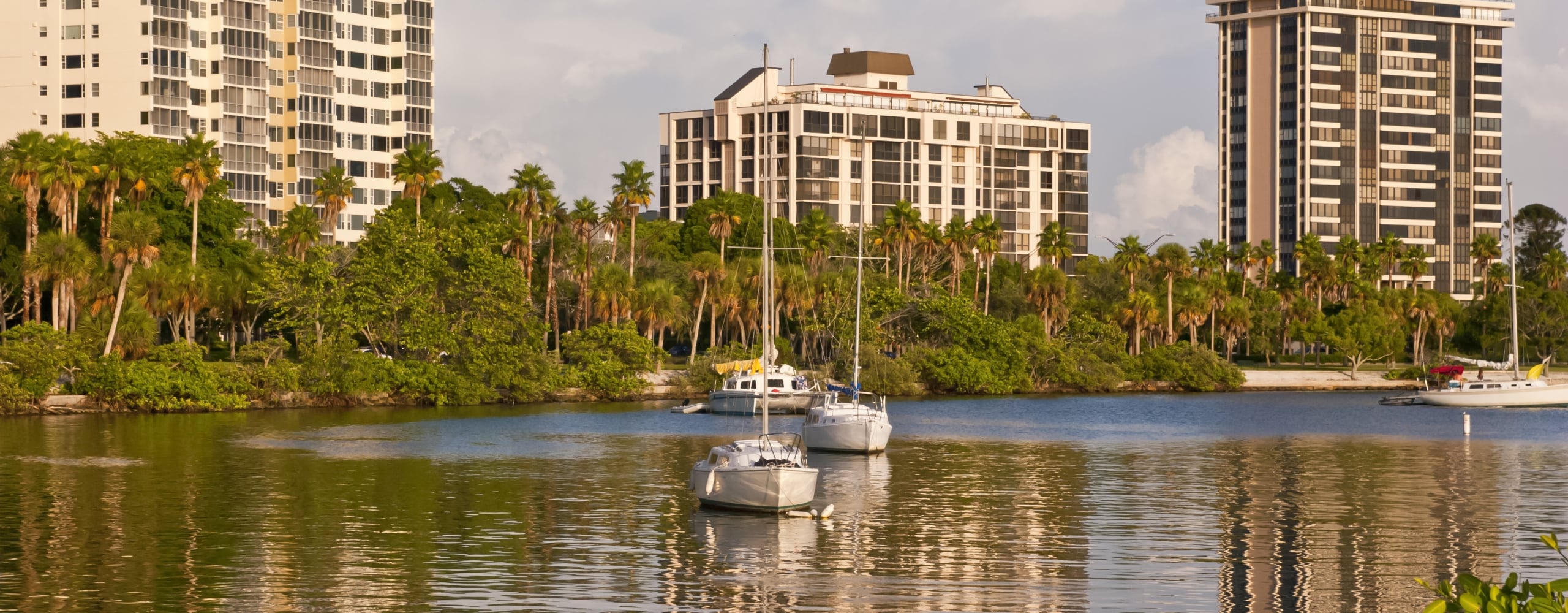 Sailboats rest on calm water reflecting tall white and dark high-rise apartment buildings nestled behind lush tropical palm trees under a partly cloudy sky.