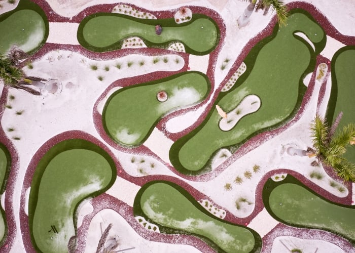 Aerial view of a tropical miniature golf course featuring irregular green putting surfaces surrounded by white sand and maroon borders, with palm trees interspersed.