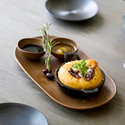 Baked cheese appetizer served on a wooden platter with dipping oils, olives, rosemary, and glasses of white and red wine on a light wood table.