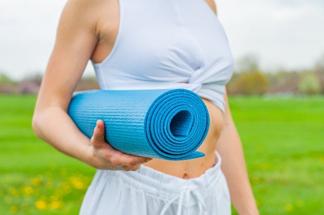 Woman in white athletic wear holding a rolled-up bright blue yoga or exercise mat outdoors in a grassy field.