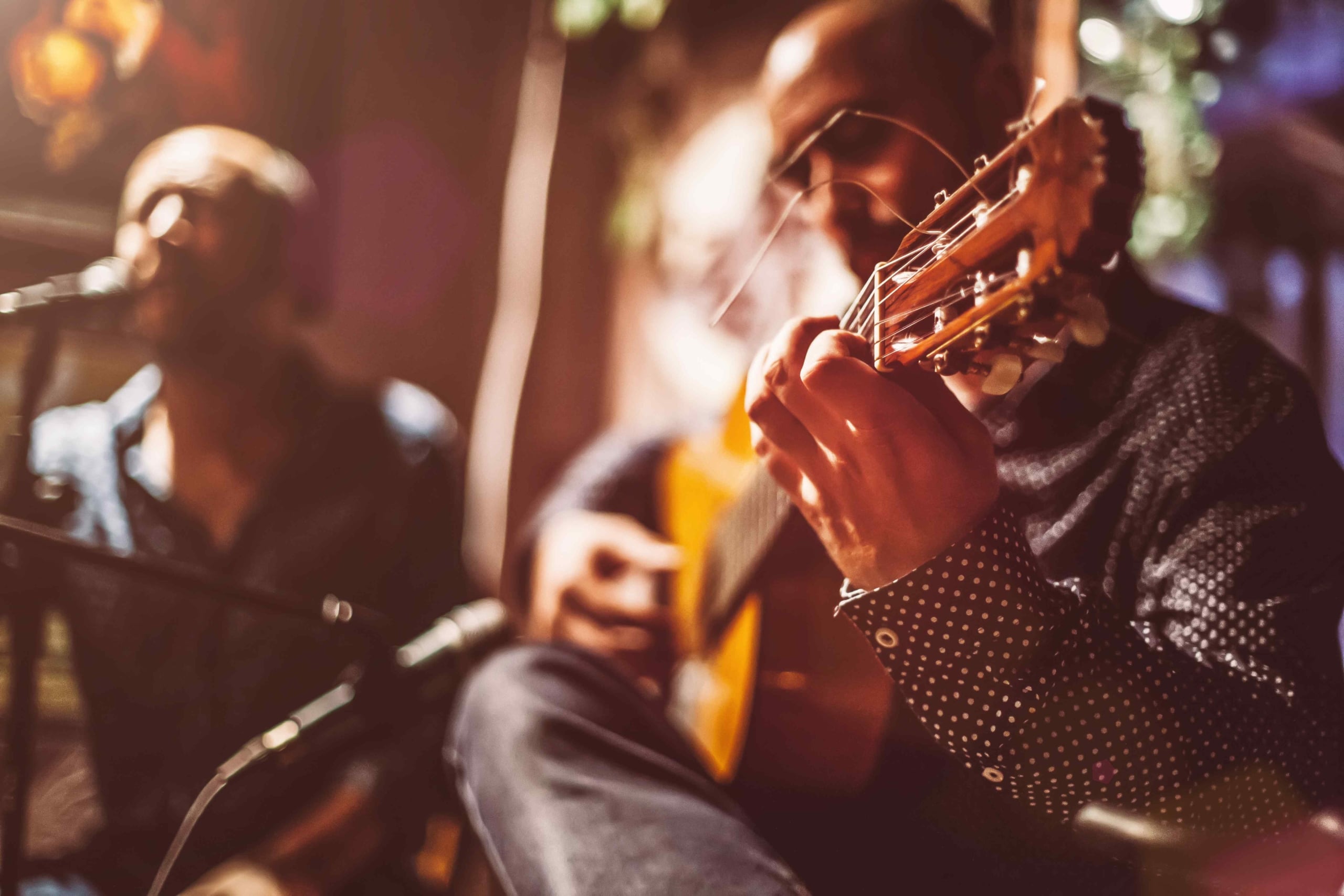 Close-up of a musician wearing a polka-dot shirt intently playing the neck of an acoustic guitar during a dimly lit live performance with another musician blurred in the background.