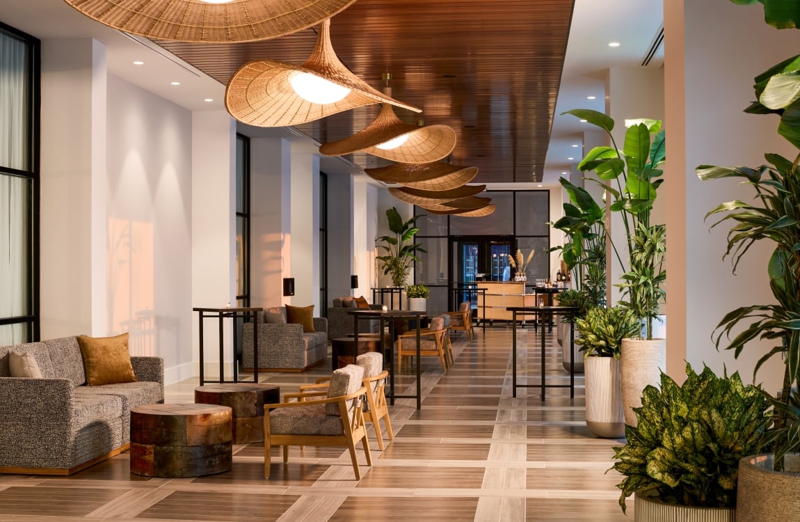 Modern hotel lobby lounge featuring woven pendant lights, rich wood accents, patterned seating, and large potted tropical plants.