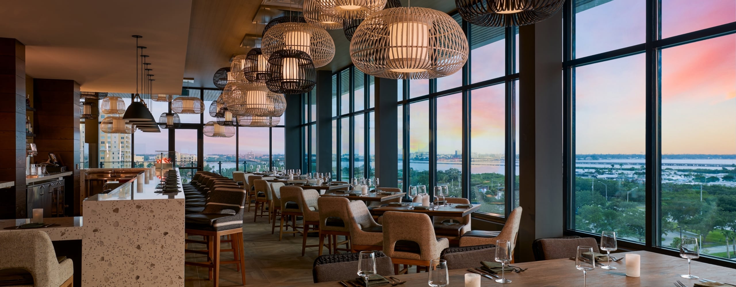 Upscale restaurant dining room featuring woven pendant lights, a speckled bar, and large windows showcasing a vibrant sunset over a cityscape and body of water.