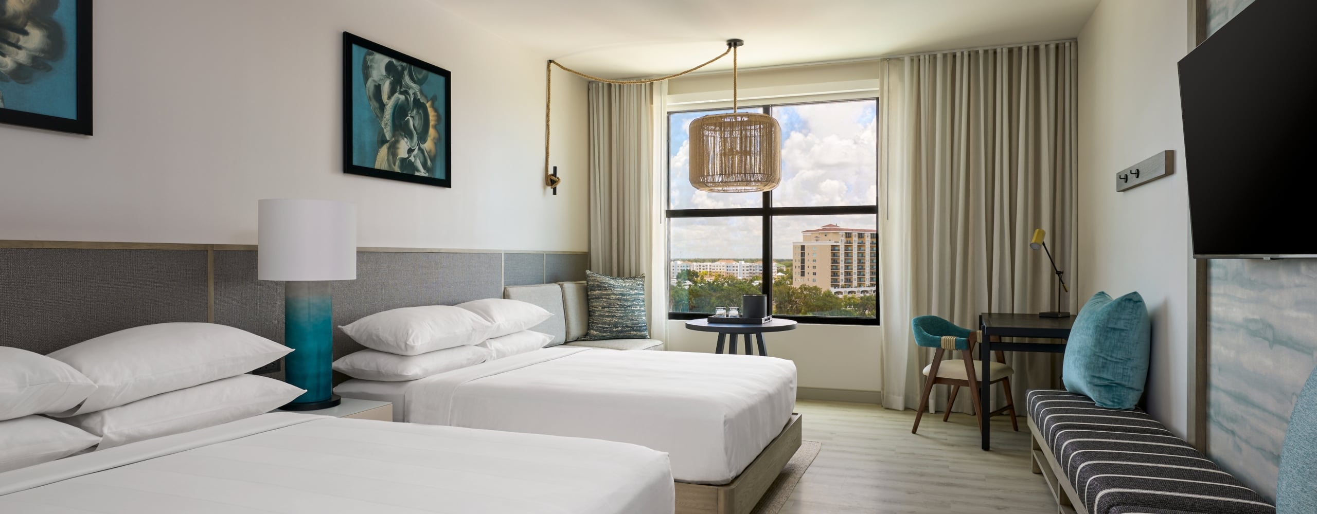 Palmetto hotel room with two double beds, a bench, a desk and chair by the window, wall art, a TV, and neutral modern decor.