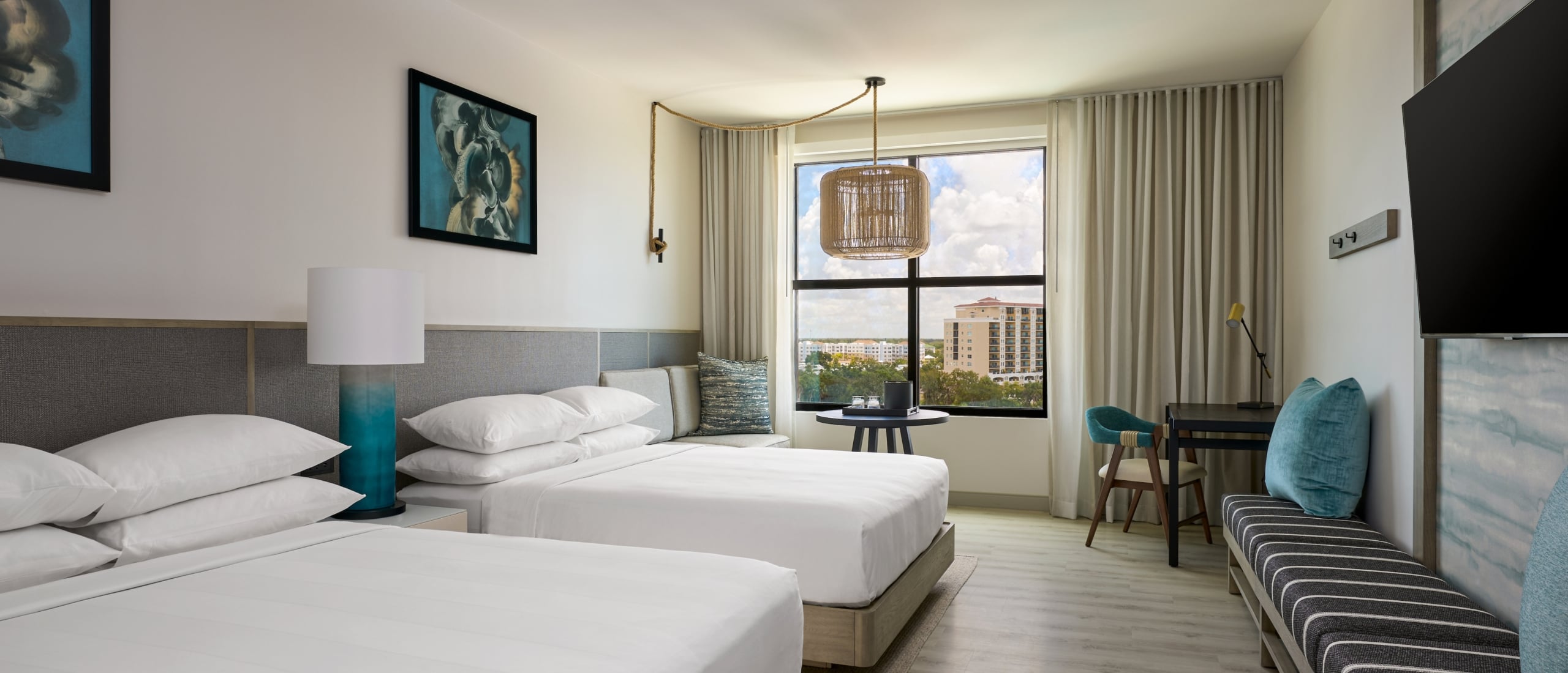 Palmetto hotel room with two double beds, a bench, a desk and chair by the window, wall art, a TV, and neutral modern decor.