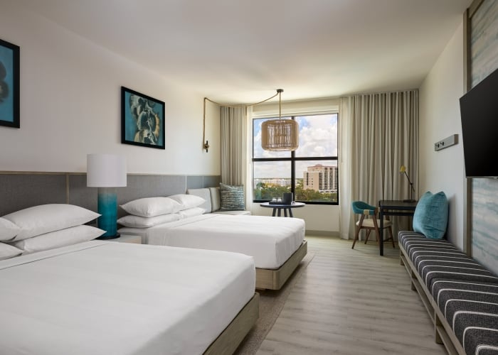 Palmetto hotel room with two double beds, a bench, a desk and chair by the window, wall art, a TV, and neutral modern decor.