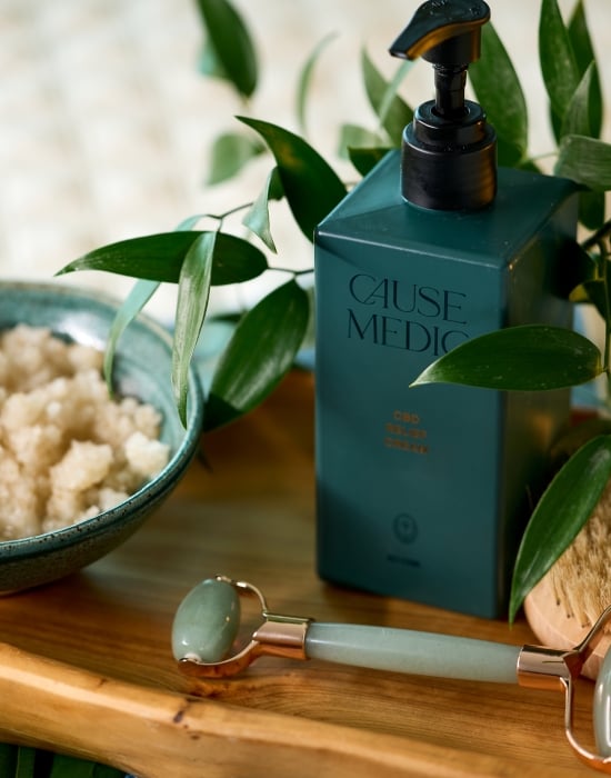 A close-up shot of skincare products, including a jar of scrub, a green pump bottle, a jade roller, and a dry brush, arranged on a wooden tray with green foliage.