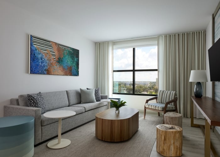 Modern living room in suite with a gray sofa, coffee tables, accent chair, wall art, floor lamp, TV, and large window with curtains letting in natural light.
