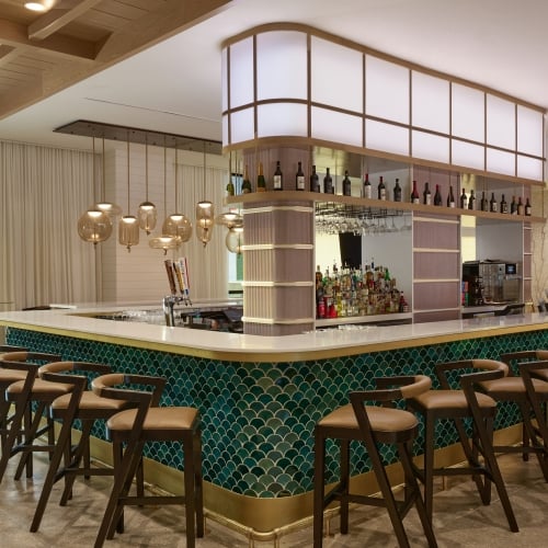 Modern hotel bar featuring a curved counter with green scallop tiles, brown leather bar stools, and overhead illuminated shelving displaying liquor bottles.