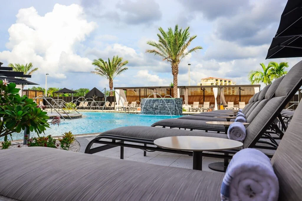 Row of modern gray chaise lounges with rolled white towels overlooks a resort swimming pool featuring a stone waterfall and palm trees under a cloudy sky.