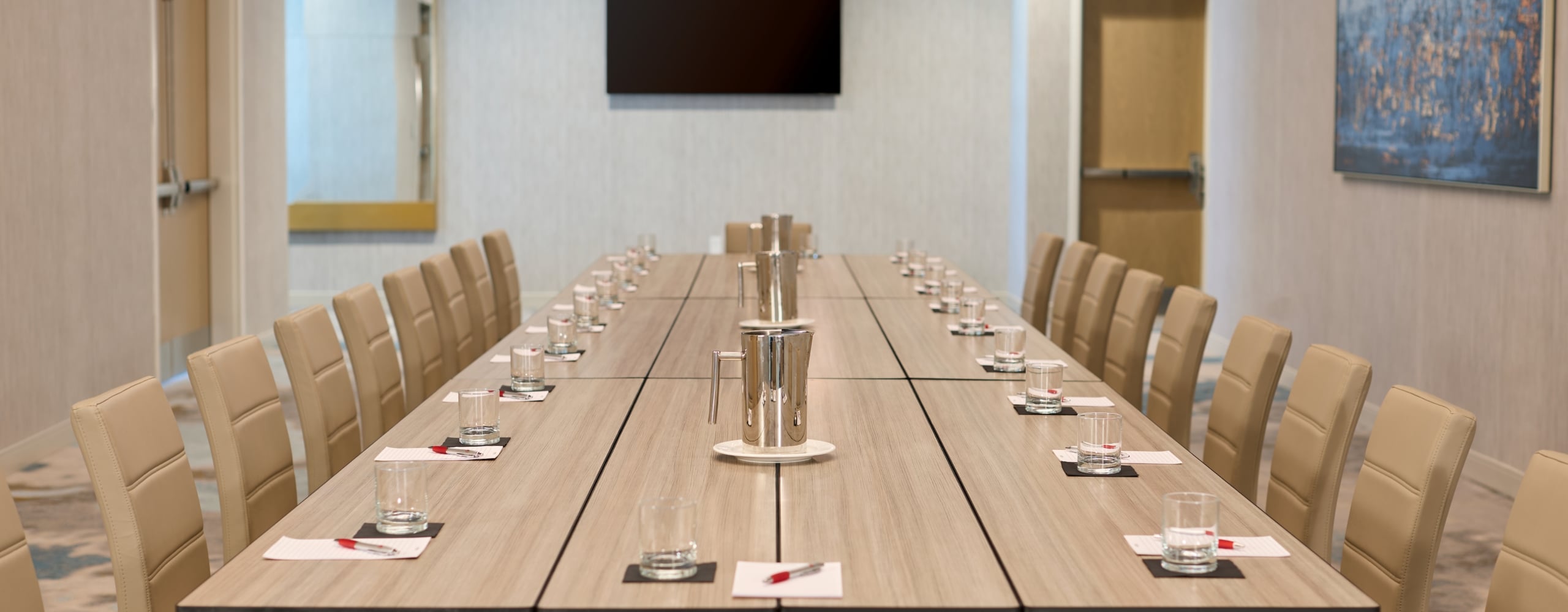Long rectangular conference table set with beige chairs, water glasses, and pitchers in a bright meeting room with a mounted screen and abstract art.