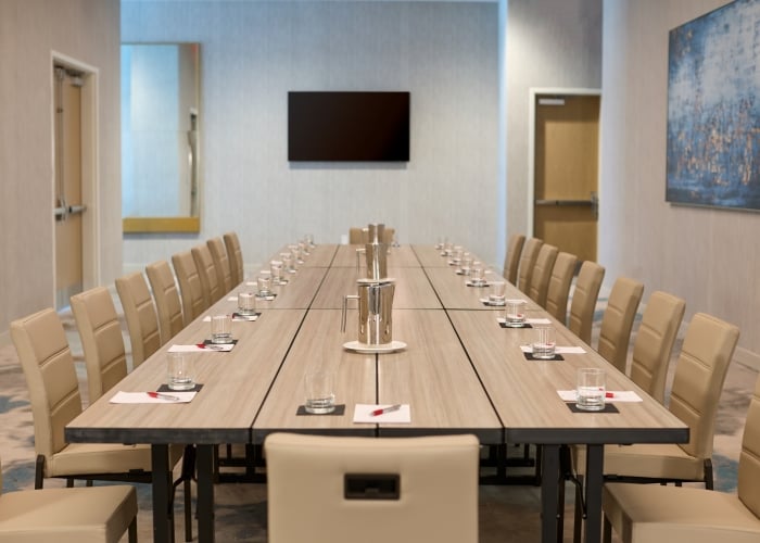 Long rectangular conference table set with beige chairs, water glasses, and pitchers in a bright meeting room with a mounted screen and abstract art.